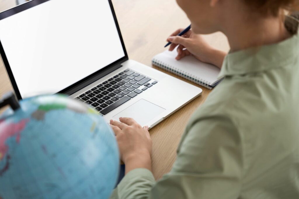Étudiant(e) apprenant en ligne avec ordinateur portable, écran blanc, cahier et globe terrestre sur un bureau.