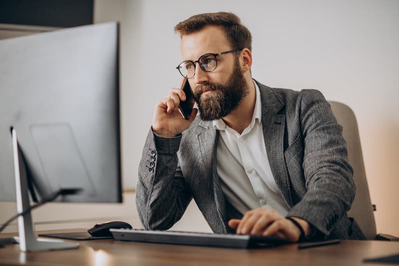 Homme d'affaires barbu en costume gris parlant au téléphone à son bureau avec ordinateur portable