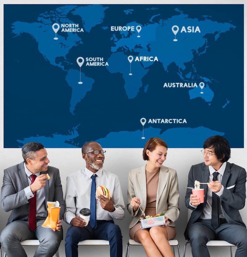 Quatre personnes en tenue professionnelle mangent de la restauration rapide tout en étant assis devant une carte du monde étiquetée avec les continents.